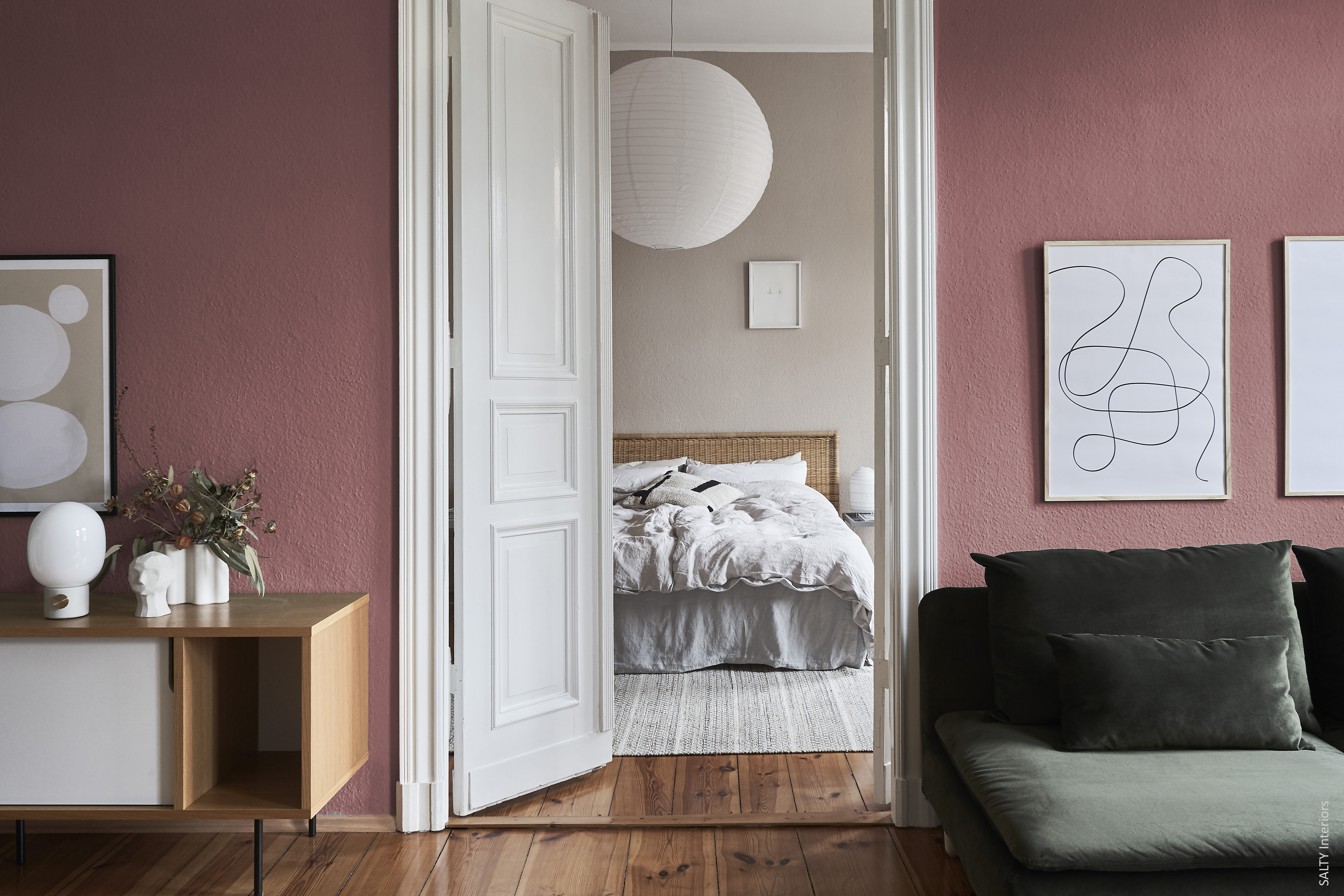 Ein gemütliches, modernes Wohnzimmer mit rosa Wänden, minimalistischer Kunst und einem grünen Sofa, das durch offene weiße Doppeltüren zu einem sanft beleuchteten Schlafzimmer mit einem Kopfteil aus Korbgeflecht und weißer Bettwäsche führt.